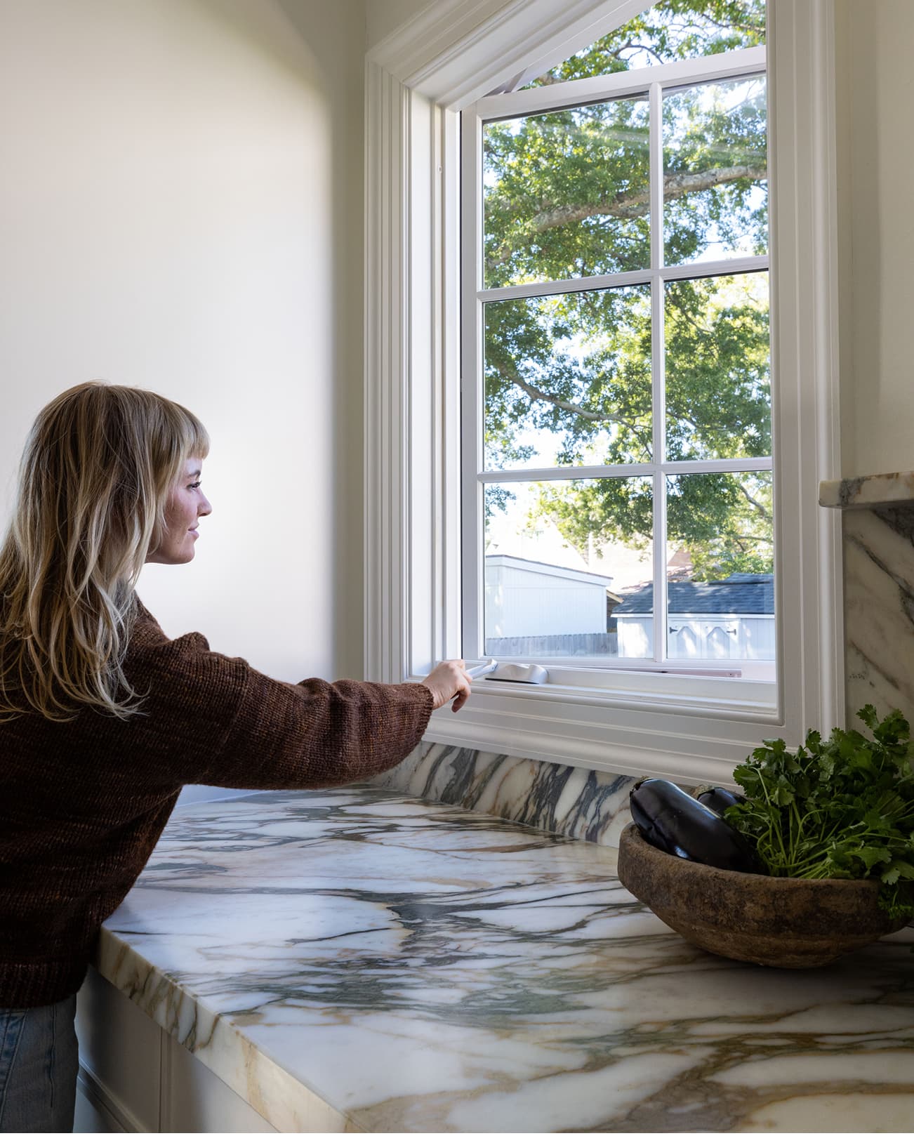 A woman reaches over a granite countertop to use the crank handle of a white Marvin Elevate Casement window with divided lites.