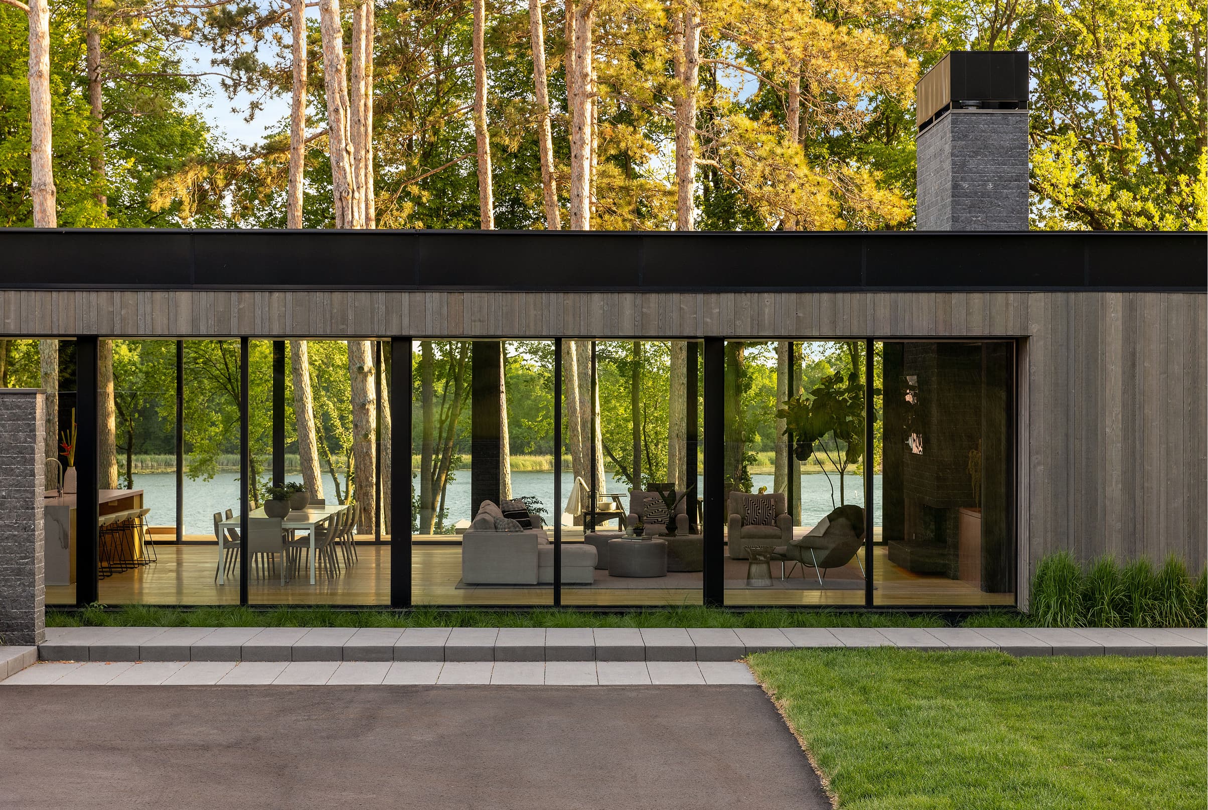 Modern home exterior with a row of large black Marvin Modern windows leading to a view of a lake and birch trees.